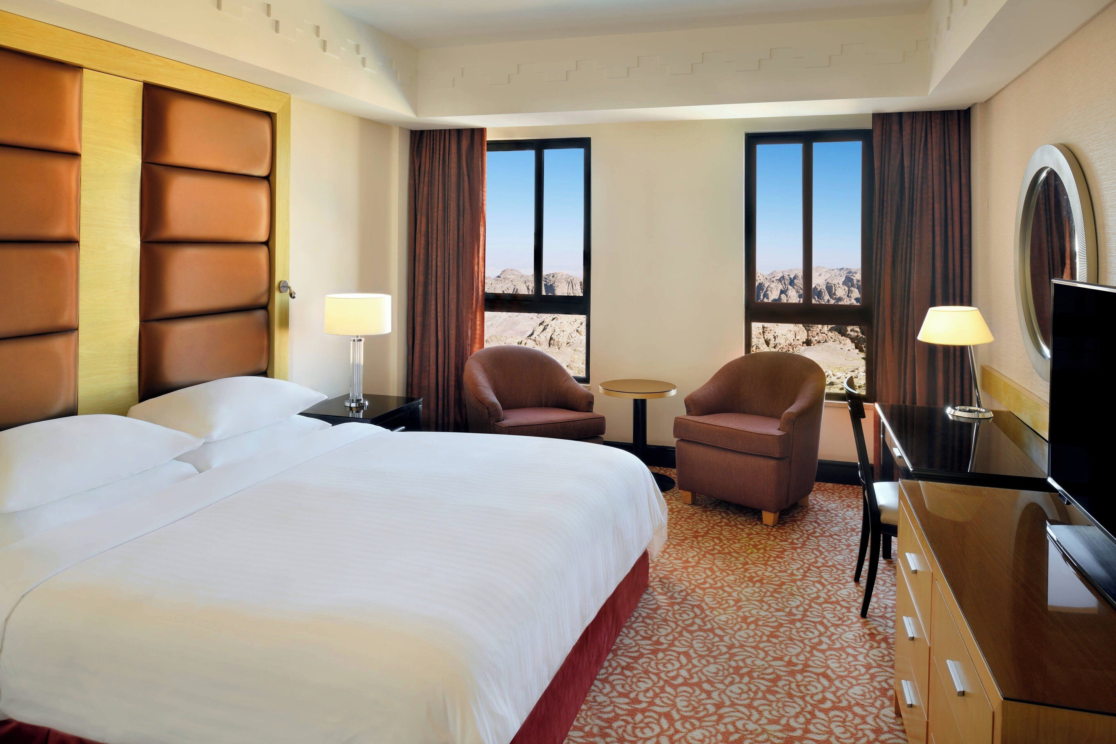 A hotel room with a large bed, two armchairs, a small table, a TV, and a view of a mountainous landscape through the windows.