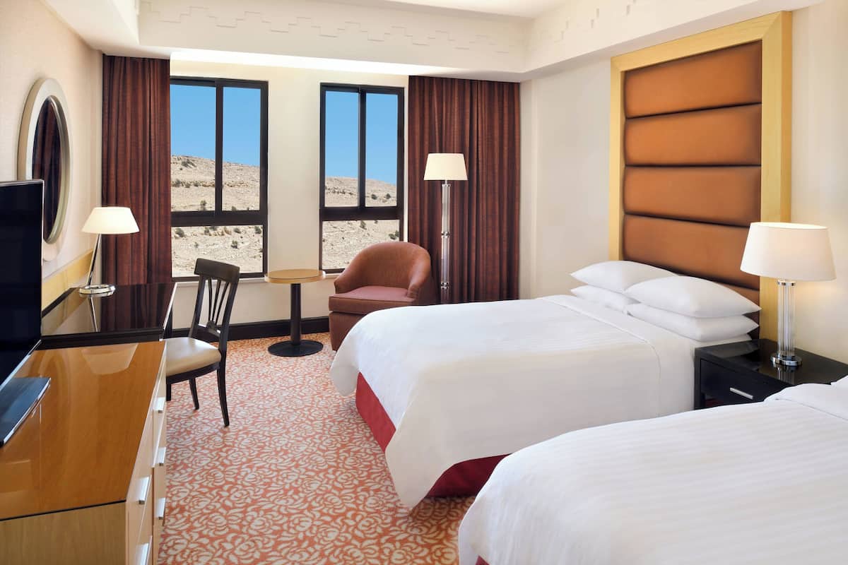 A hotel room with two beds, a desk, a chair, a TV, and a view of a desert landscape.