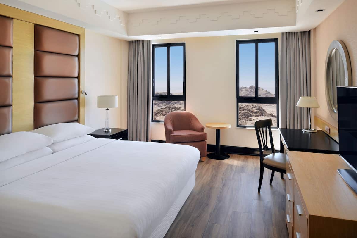 A hotel room with a large bed, a desk with a chair, a TV, and a view of the desert through the windows.