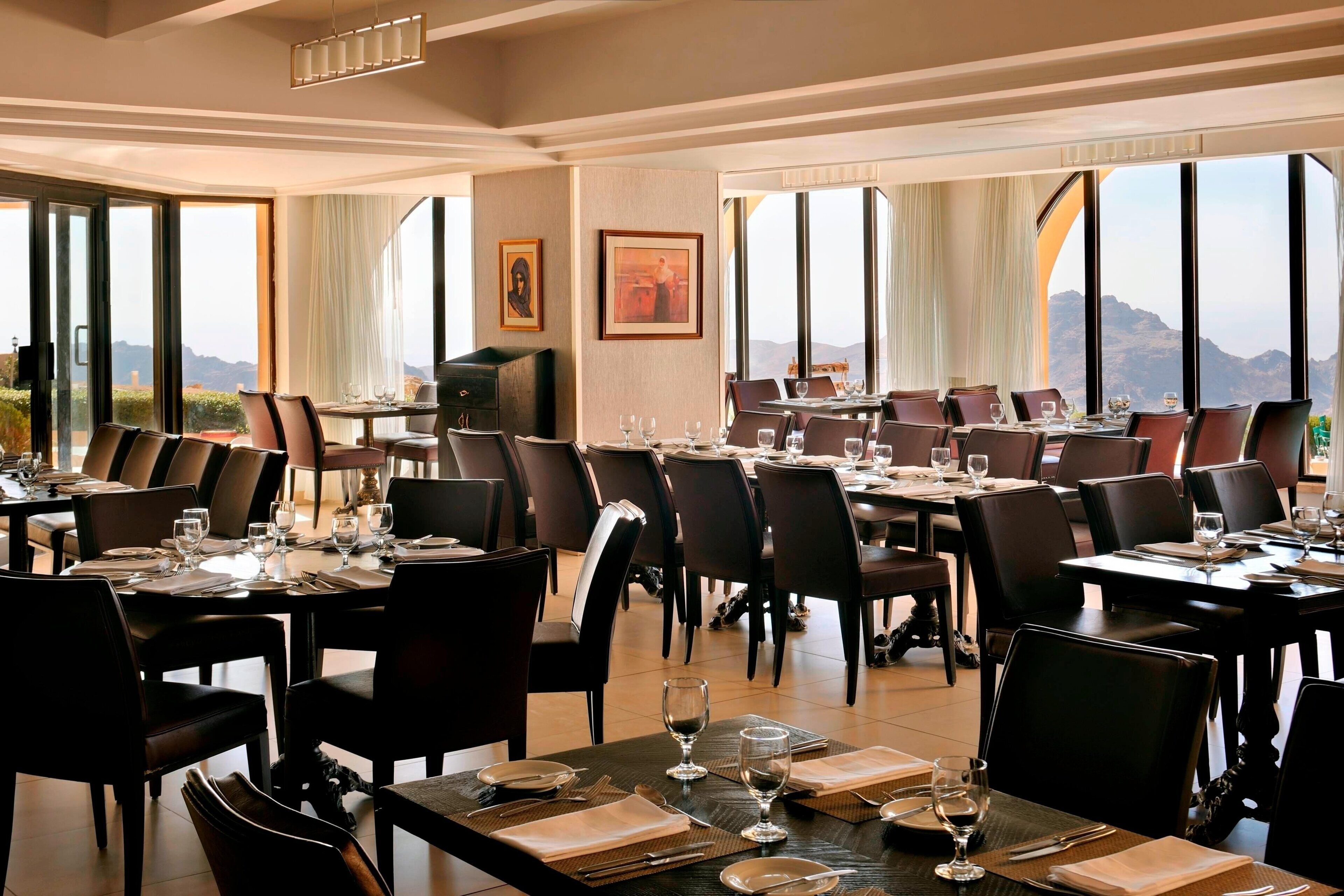 A dining area with multiple tables and chairs, large windows with mountain views, and framed artwork on the walls.