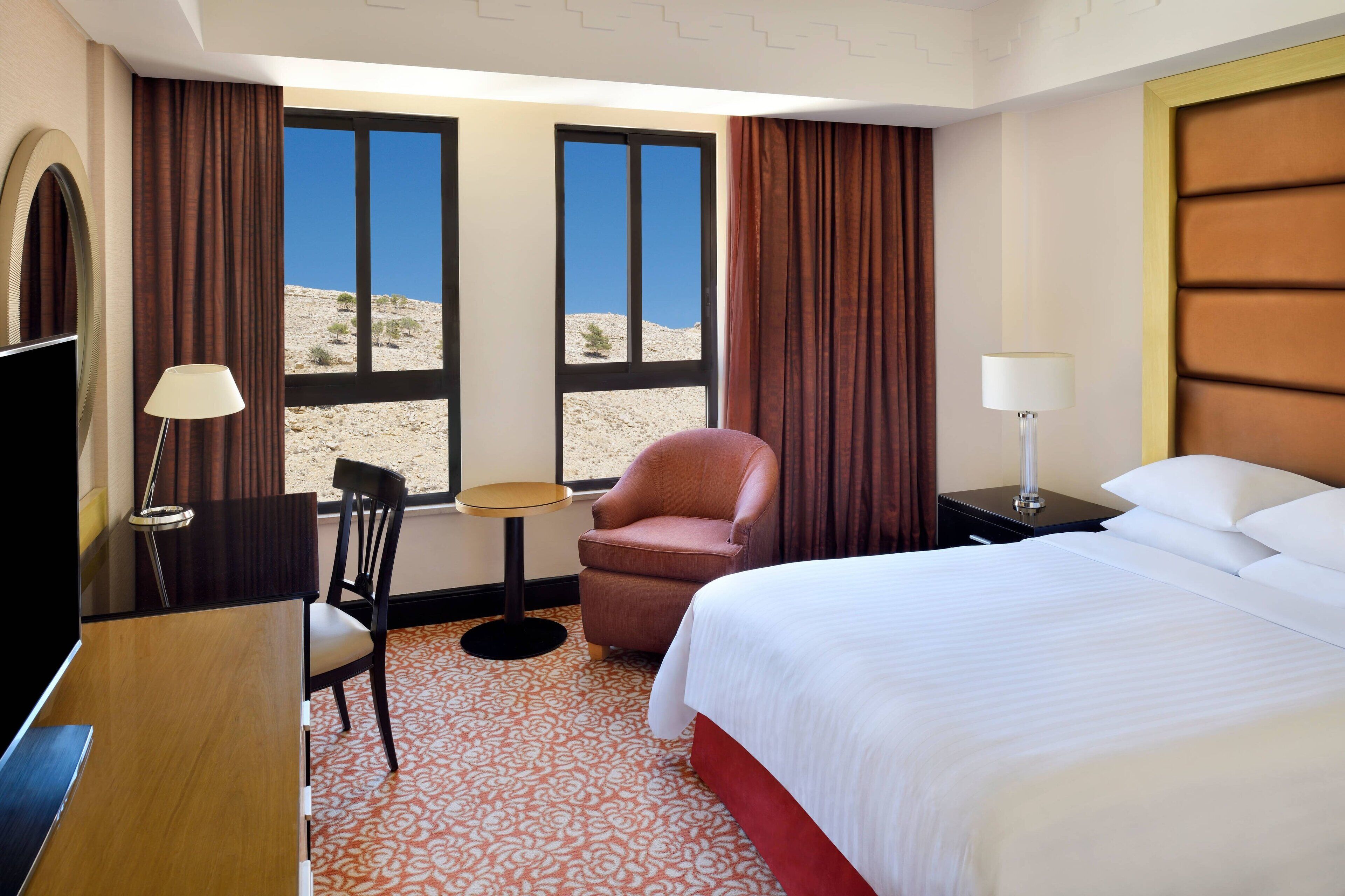 Habitación de hotel con cama, escritorio, silla, lámpara y vista a un paisaje desértico a través de ventanas amplias.