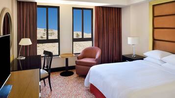 A hotel room with a bed, a desk, a chair, a lamp, and a view of a desert landscape through large windows.