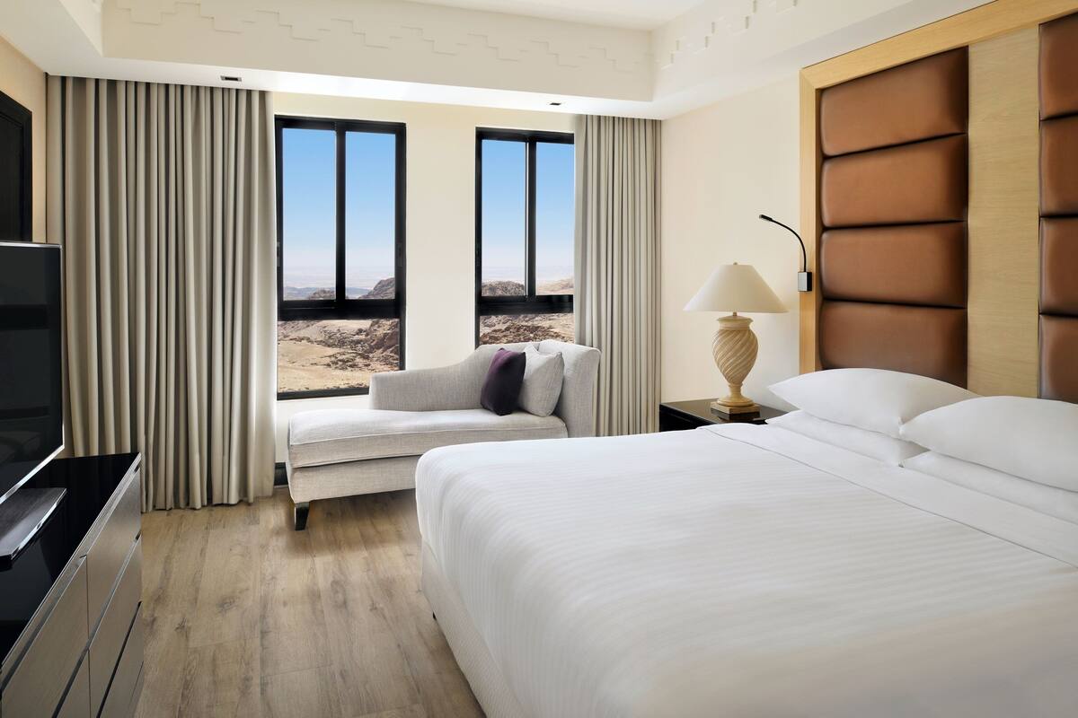 A hotel room with a bed, a sofa, a TV, a lamp, and a view of a desert landscape through large windows.