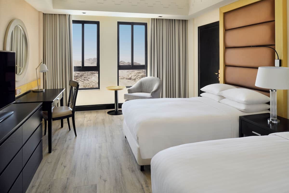 A hotel room with two beds, a desk, a chair, and a view of the desert through large windows.