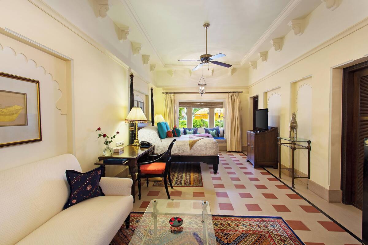 A hotel room with a sofa, a coffee table, a bed with colorful bedding, a desk with a lamp, and a television.