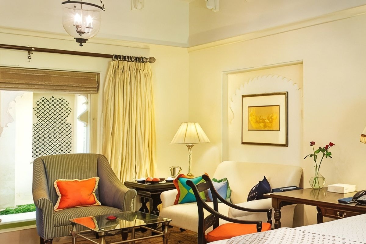 A hotel room with a bed, two armchairs, a small table, a nightstand, a lamp, a vase with flowers, and a window with curtains.