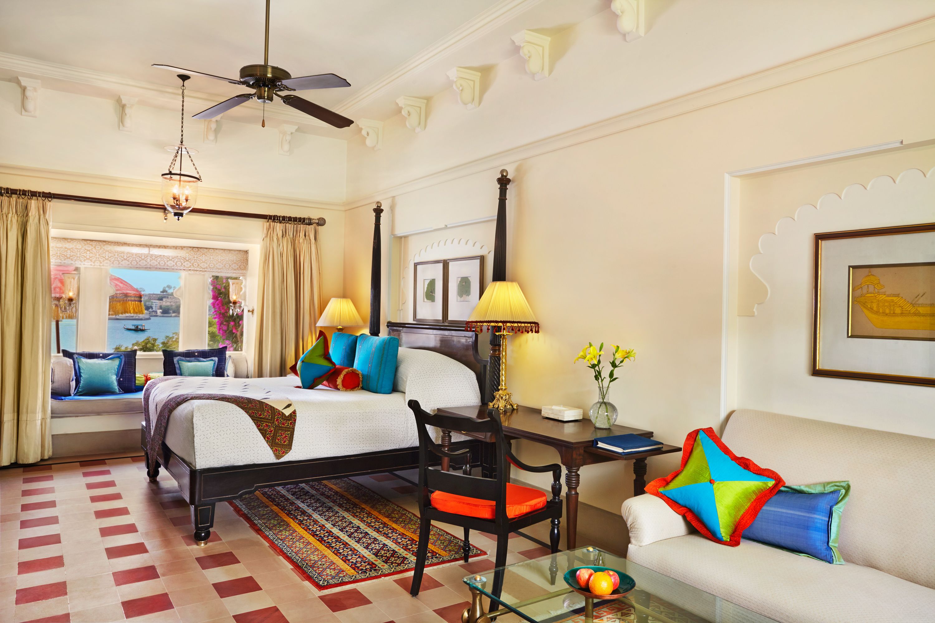 A hotel room with a bed, two chairs, a glass table, a desk, a ceiling fan, and a view of the outdoors.