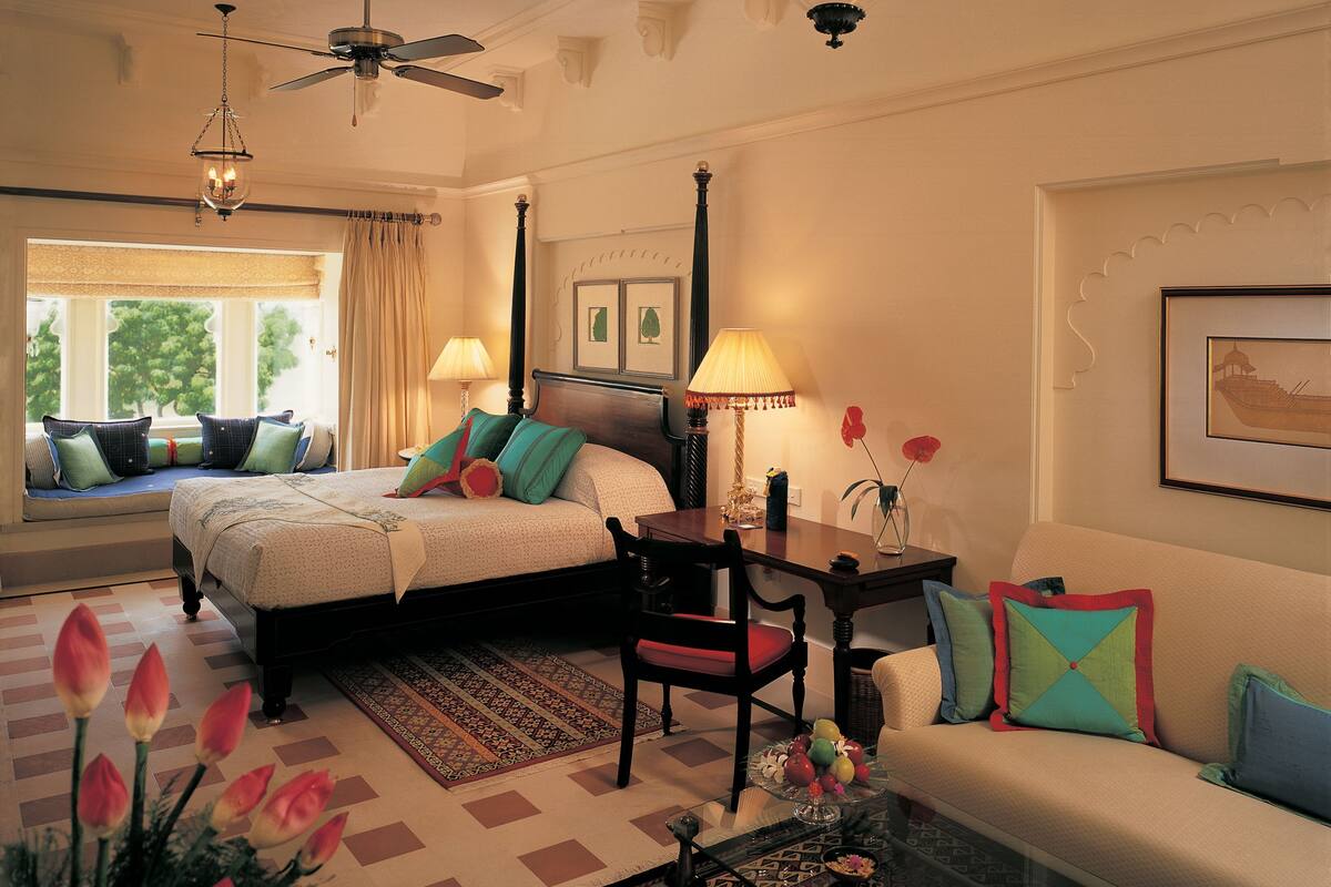 A hotel room with a bed, a sofa, a desk, and a chair. There is a window with curtains, a chandelier, and a framed picture on the wall.