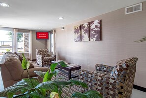 A hotel lobby with a seating area, a television displaying 'Free Wi-Fi All Rooms,' and a large window with a view of palm trees.