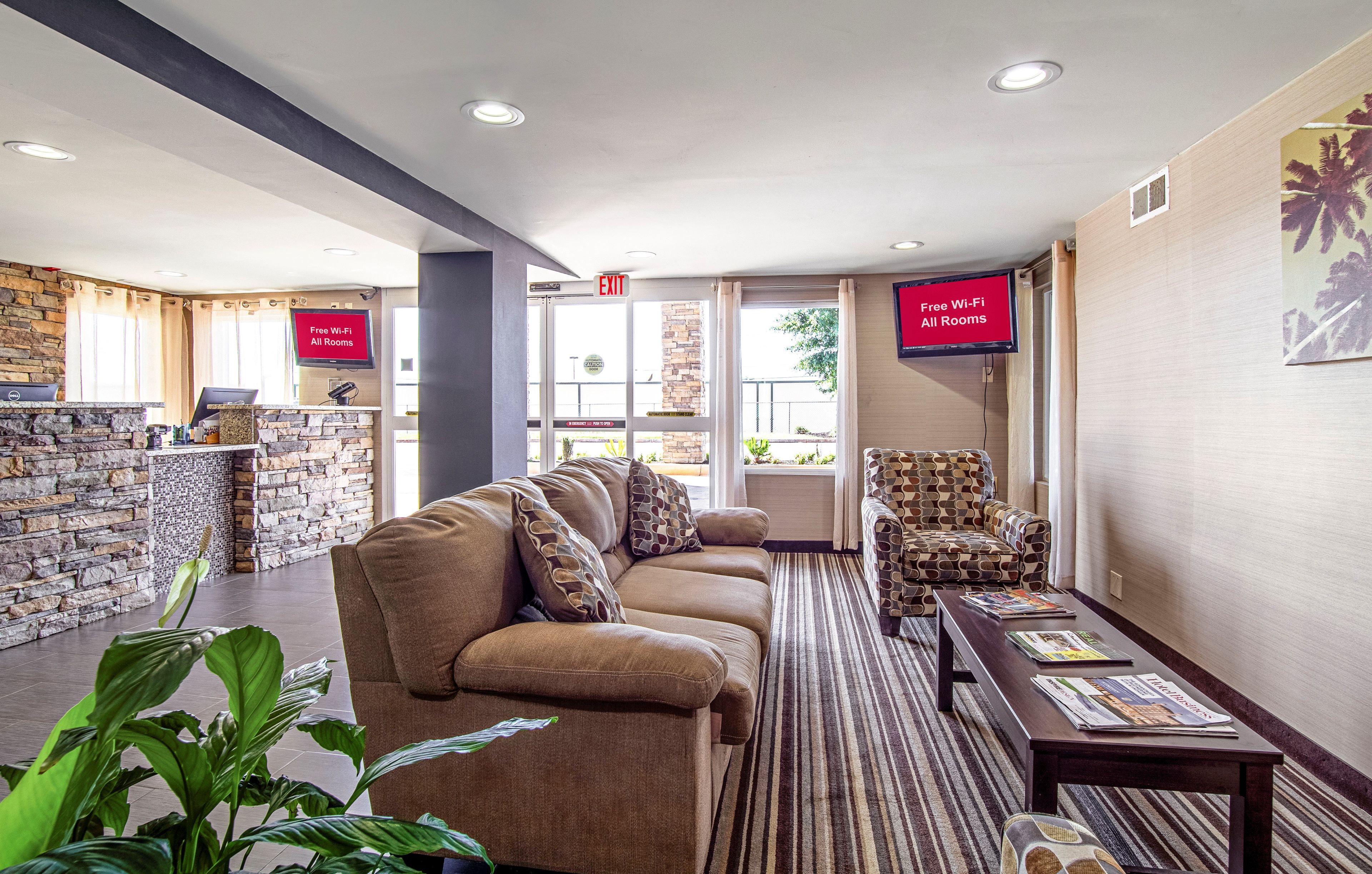A lobby with a stone wall, a reception desk, a sofa, a patterned armchair, and a television screen displaying 'Free Wi-Fi All Rooms'.