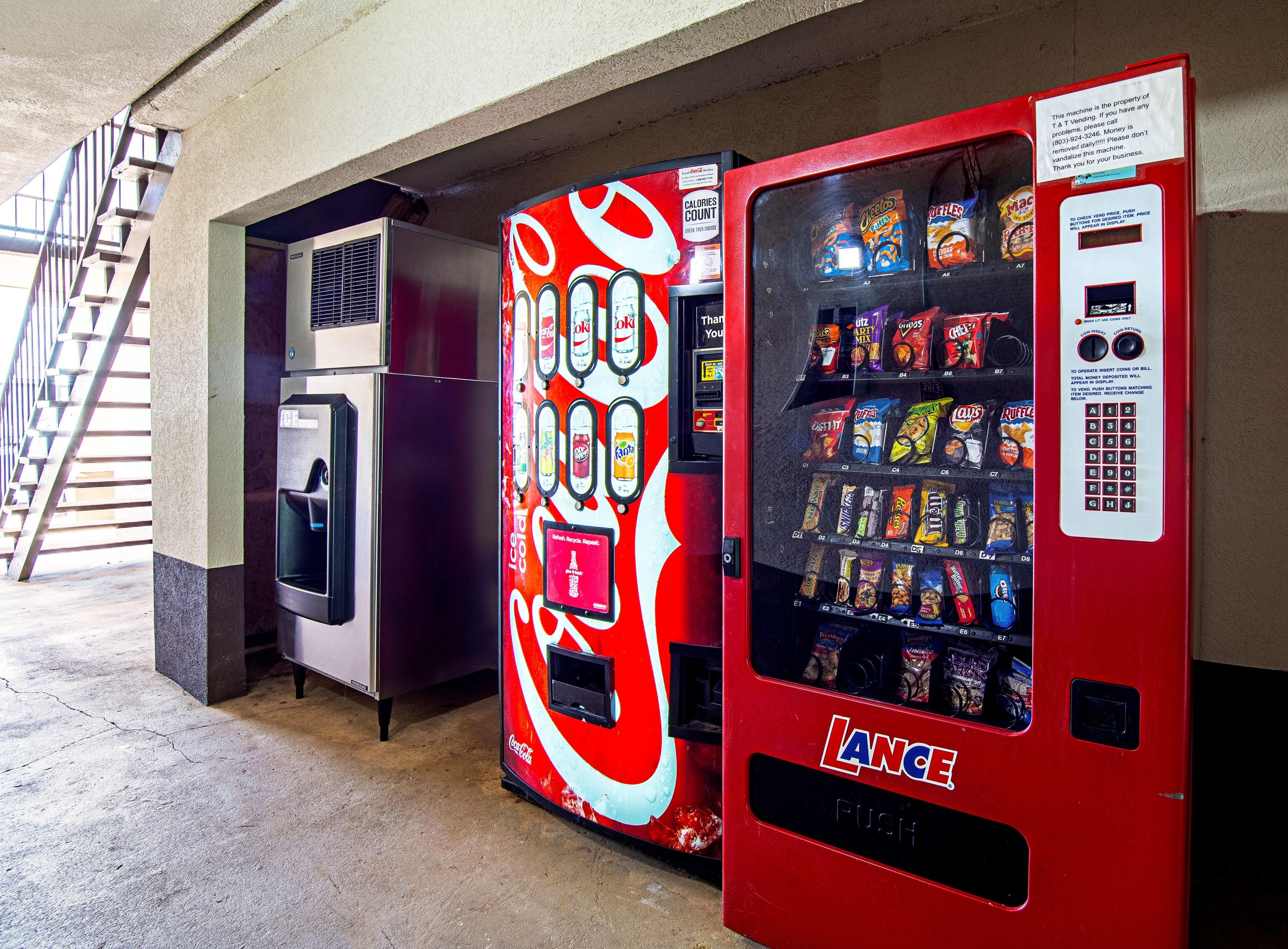 Máquina de vendas com exterior vermelho e logótipo da Coca-Cola, situada num espaço interior com chão de betão e escadas ao fundo.
