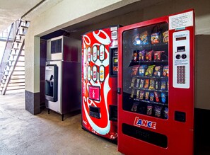 Un distributeur automatique à l’extérieur rouge, orné du logo Coca-Cola, situé dans un espace intérieur avec un sol en béton et un escalier en arrière-plan.