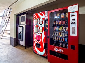 Máquina expendedora con exterior rojo y el logotipo de Coca-Cola, ubicada en un espacio interior con piso de concreto y una escalera al fondo.