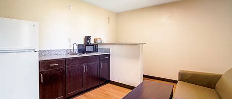 A compact kitchen with dark cabinets, a white refrigerator, and a microwave on the countertop. There's a small table and a beige sofa in the living area.