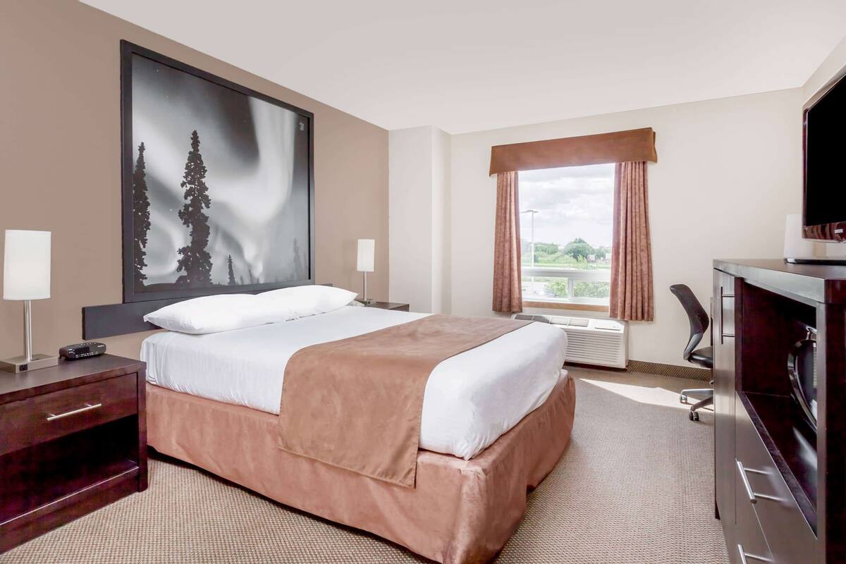 A hotel room with a bed, a desk with a computer, a TV, a window with curtains, and a large painting on the wall.