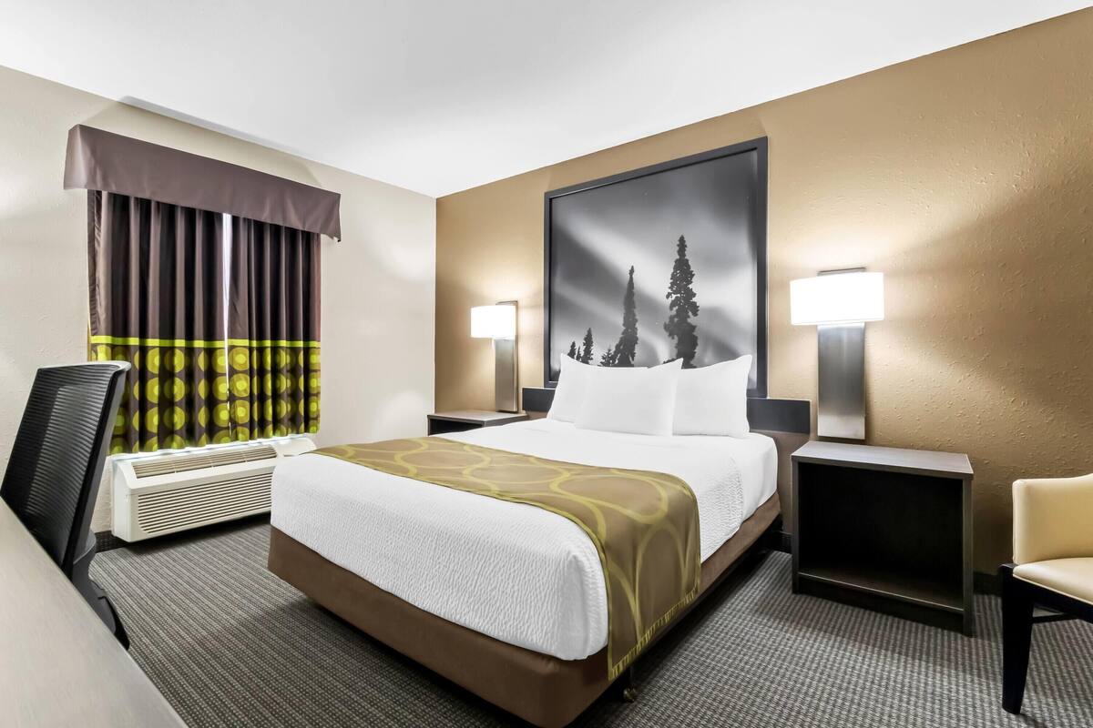 A hotel room with a large bed, a desk, a chair, a desk lamp, a wall-mounted TV, and a window with curtains.