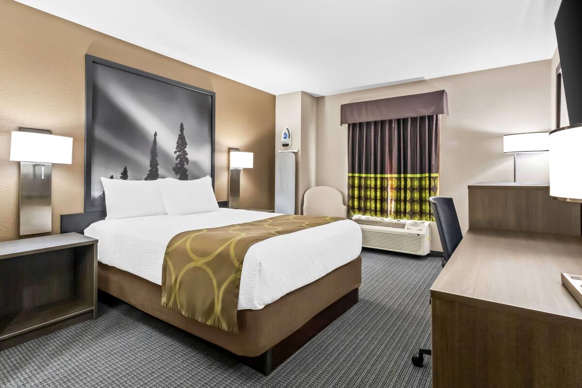 A hotel room with a large bed, a desk, two bedside lamps, a large window with curtains, and a wall-mounted air conditioning unit.