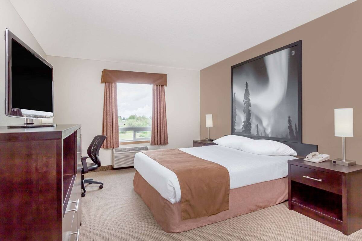 A hotel room with a bed, a desk with a TV, a chair, a window with curtains, a nightstand with a lamp, and a large painting on the wall.