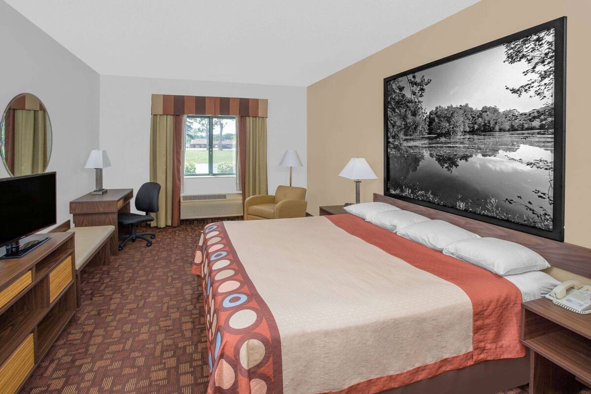Habitación de hotel con cama, escritorio con televisor, silla, lámpara de escritorio, ventana grande y un cuadro de un lago.