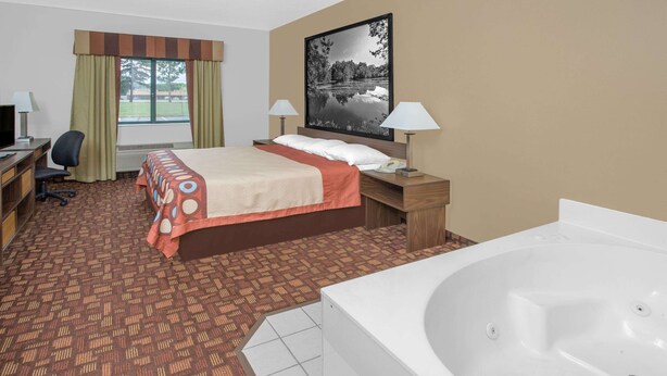 Quarto de hotel com cama, escrivaninha com computador, cadeira, abajur, mesa de cabeceira com luminária, janela grande com cortinas, quadro na parede e banheira.