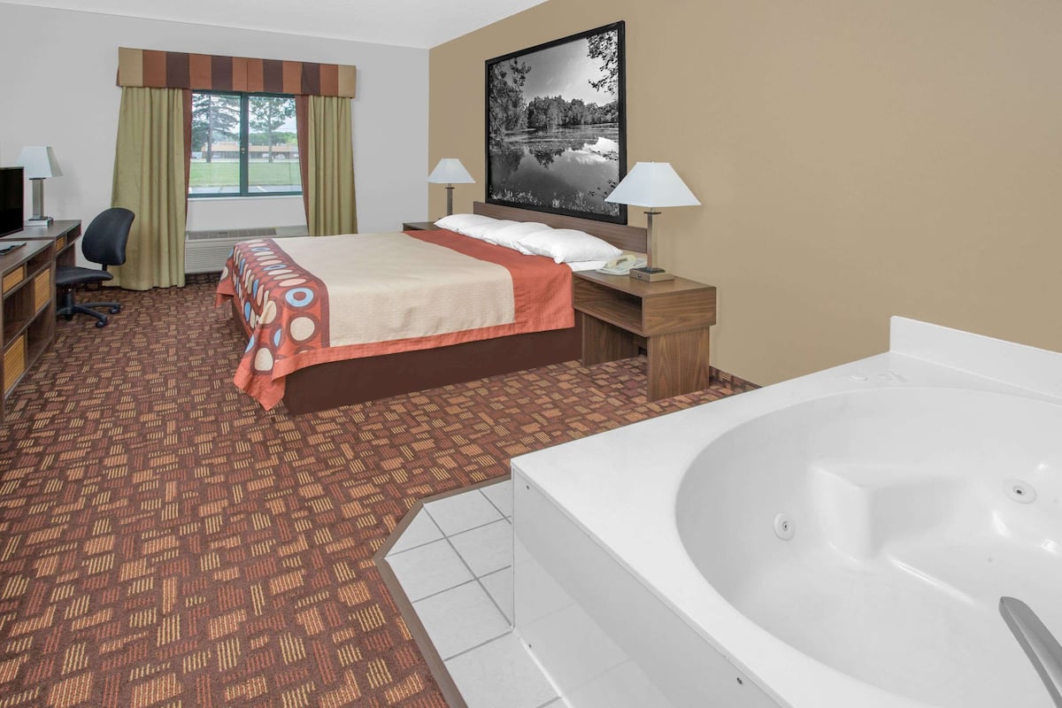 Habitación de hotel con cama, escritorio con computadora, silla, lámpara de escritorio, mesita de noche con lámpara, ventana grande con cortinas, un cuadro en la pared y una bañera.
