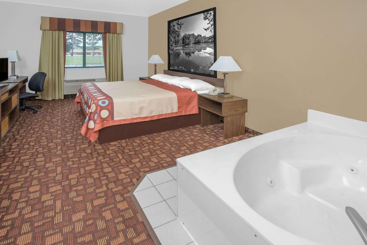 Habitación de hotel con cama, escritorio, silla, lámparas, un cuadro y una bañera.