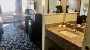 Hotellihuone, jossa on pieni keittiö, sänky, työpöytä, tuoli, televisio, kahvinkeitin, pesuallas ja roskakori.