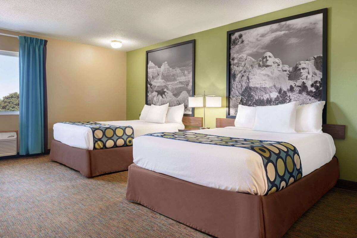 Une chambre d’hôtel avec deux lits, une fenêtre avec des rideaux, une lampe et deux grands tableaux accrochés au mur.