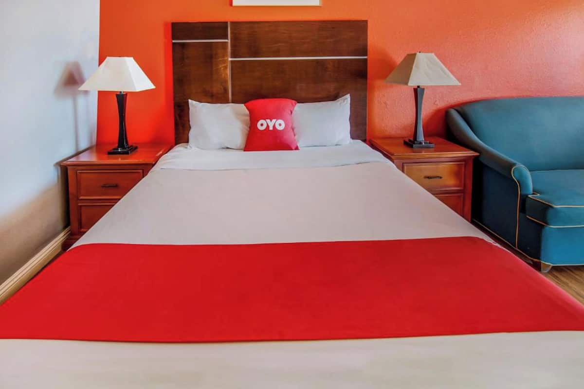 A hotel room with a large bed, two bedside tables with lamps, a blue armchair, and a red pillow with the logo "OYO".