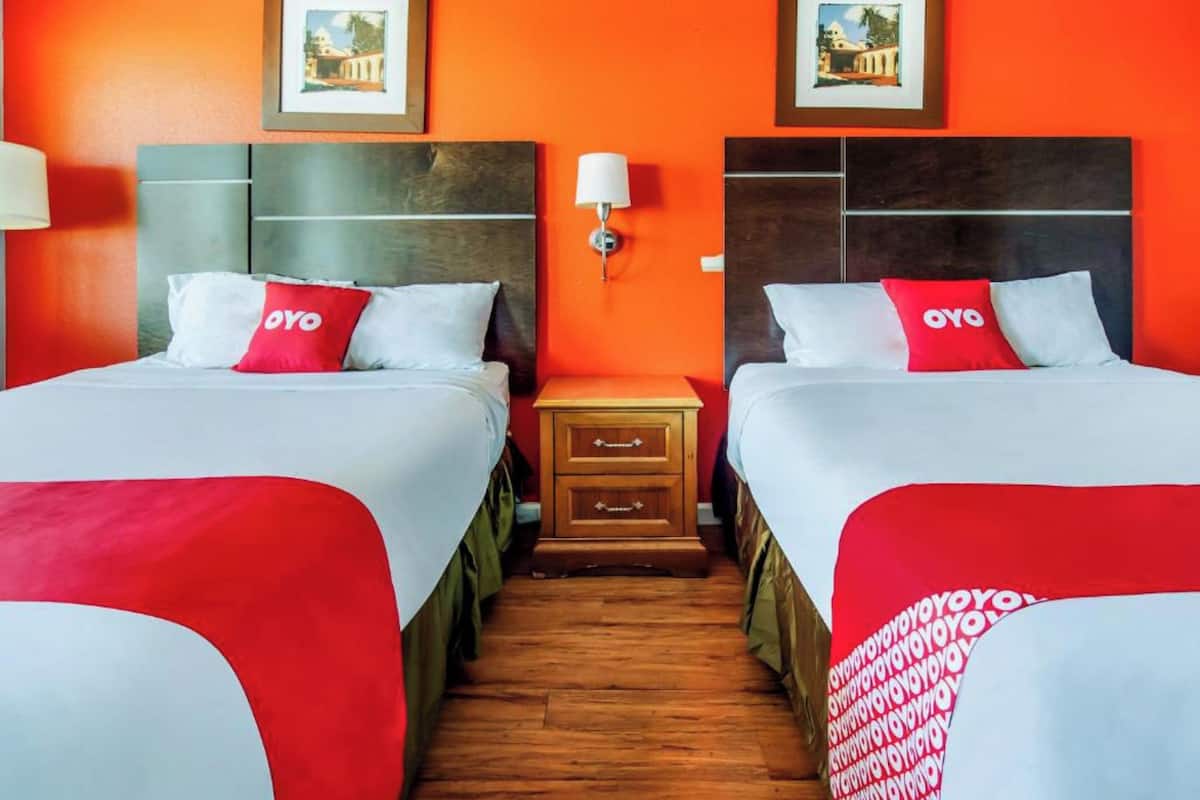 A hotel room with two beds, each with a red and white bedspread and a red pillow with "OYO" branding. There is a wooden nightstand with drawers between the beds, and a wall-mounted lamp above it. The room has orange walls and wooden flooring.