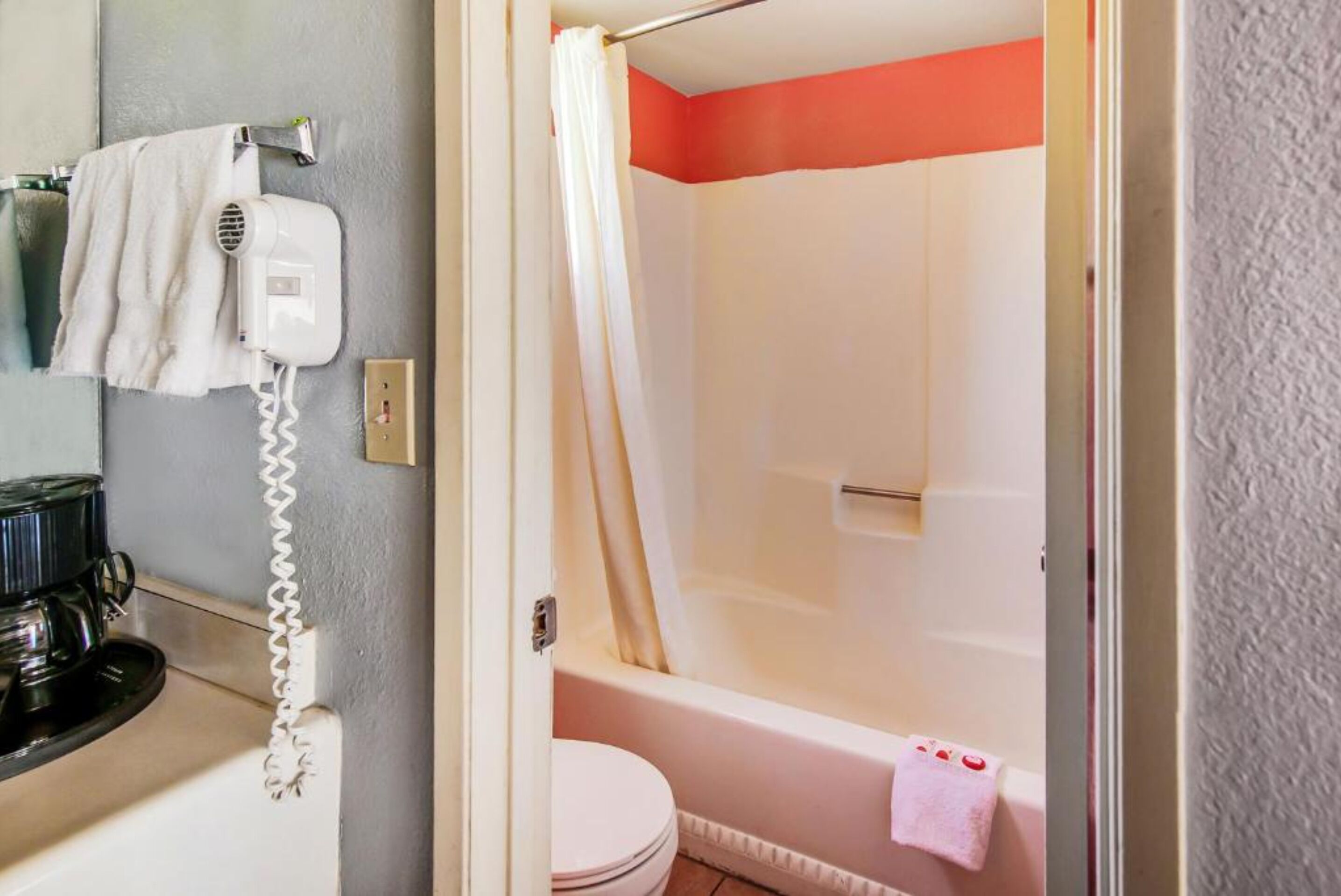 Un baño con inodoro blanco, ducha con cortina color beige y secador de pelo en la pared.