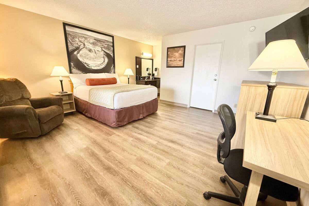 A hotel room with a bed, a desk, a chair, a lamp, and a painting on the wall.