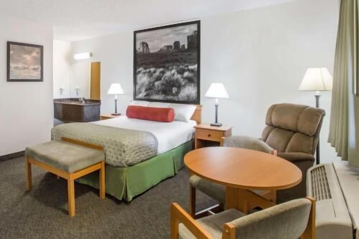 A hotel room with a bed, a desk, a chair, a round table, and a painting on the wall.