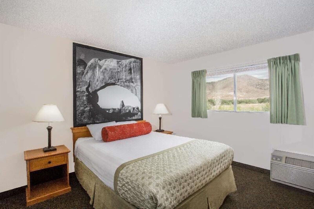 A hotel room with a bed, two bedside lamps, a large framed picture, a window with curtains, and an air conditioning unit.