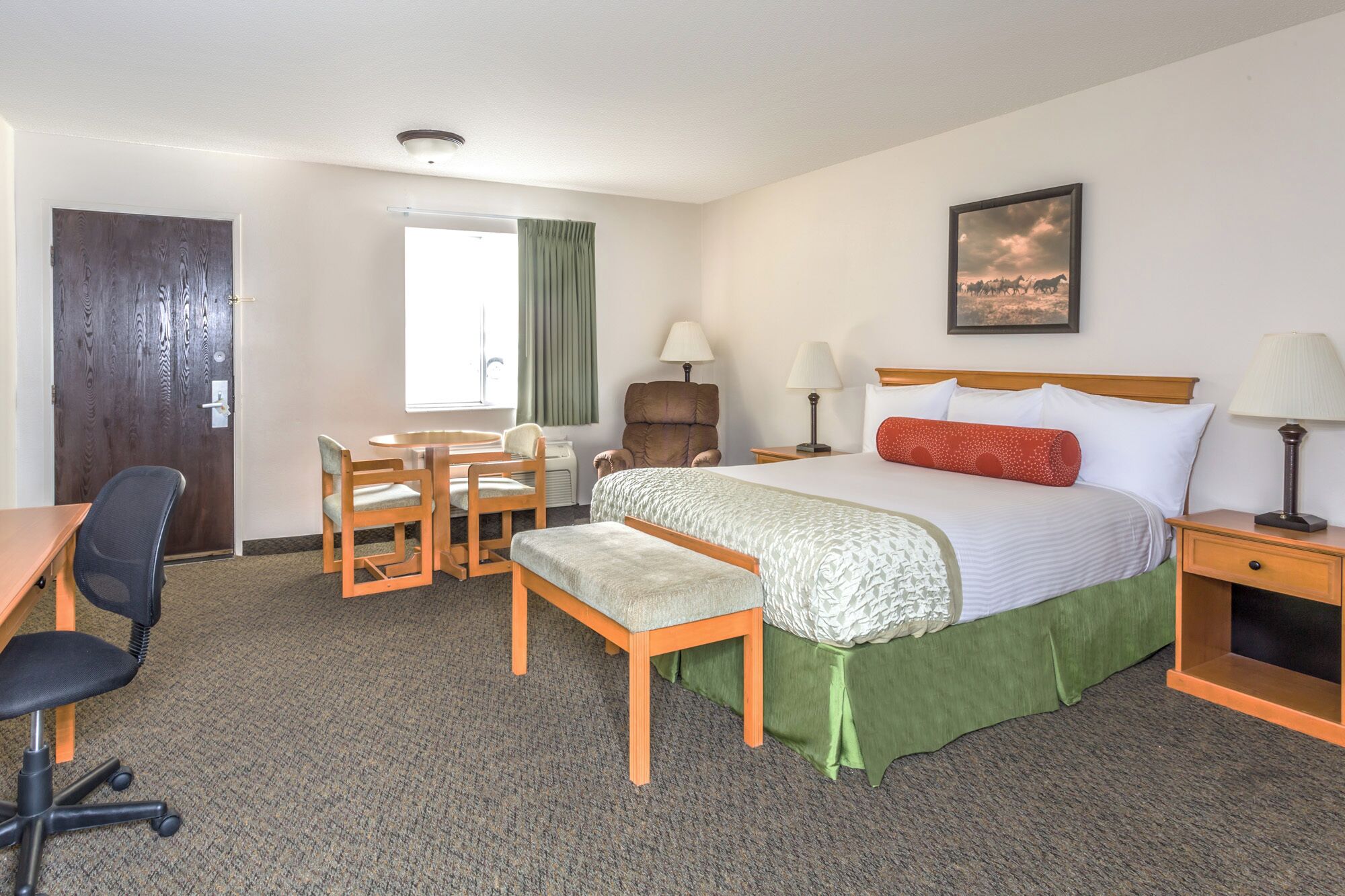 A hotel room with a bed, a desk, a chair, a small table, and a lamp.
