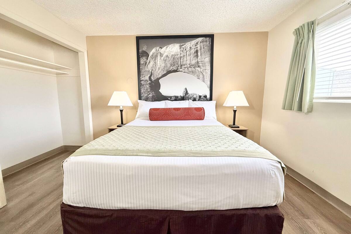 A neatly made bed with a white comforter and a red accent pillow, flanked by two bedside lamps. A large black and white photograph of a rock formation is centered above the bed.
