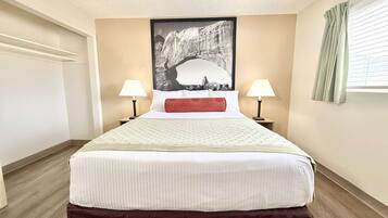 Una cama bien hecha con edredón blanco y una almohada roja decorativa, flanqueada por dos lámparas de noche. Una fotografía grande en blanco y negro de una formación rocosa está centrada sobre la cama.