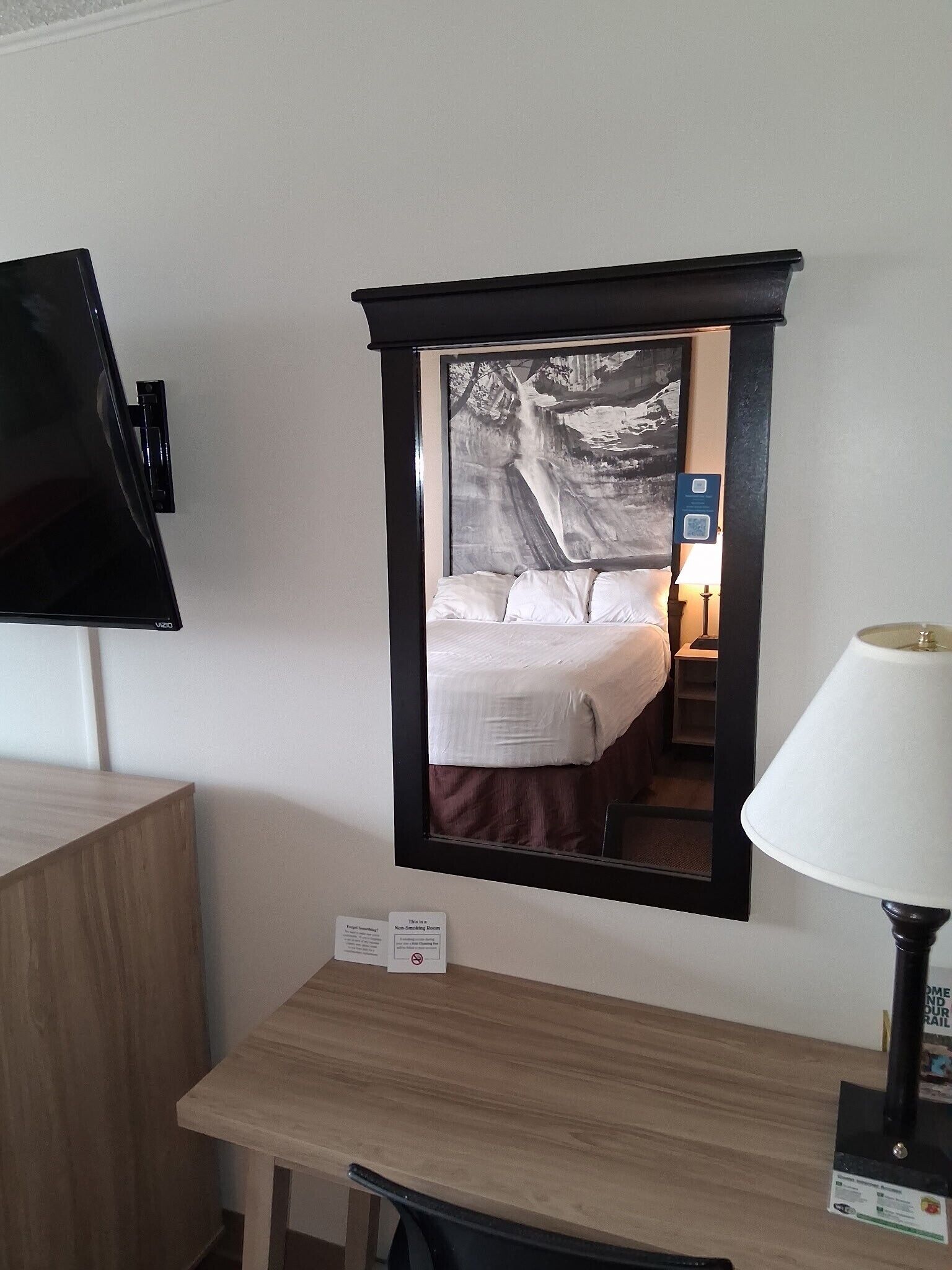 A hotel room with a bed, a desk, a lamp, and a mirror reflecting the room.