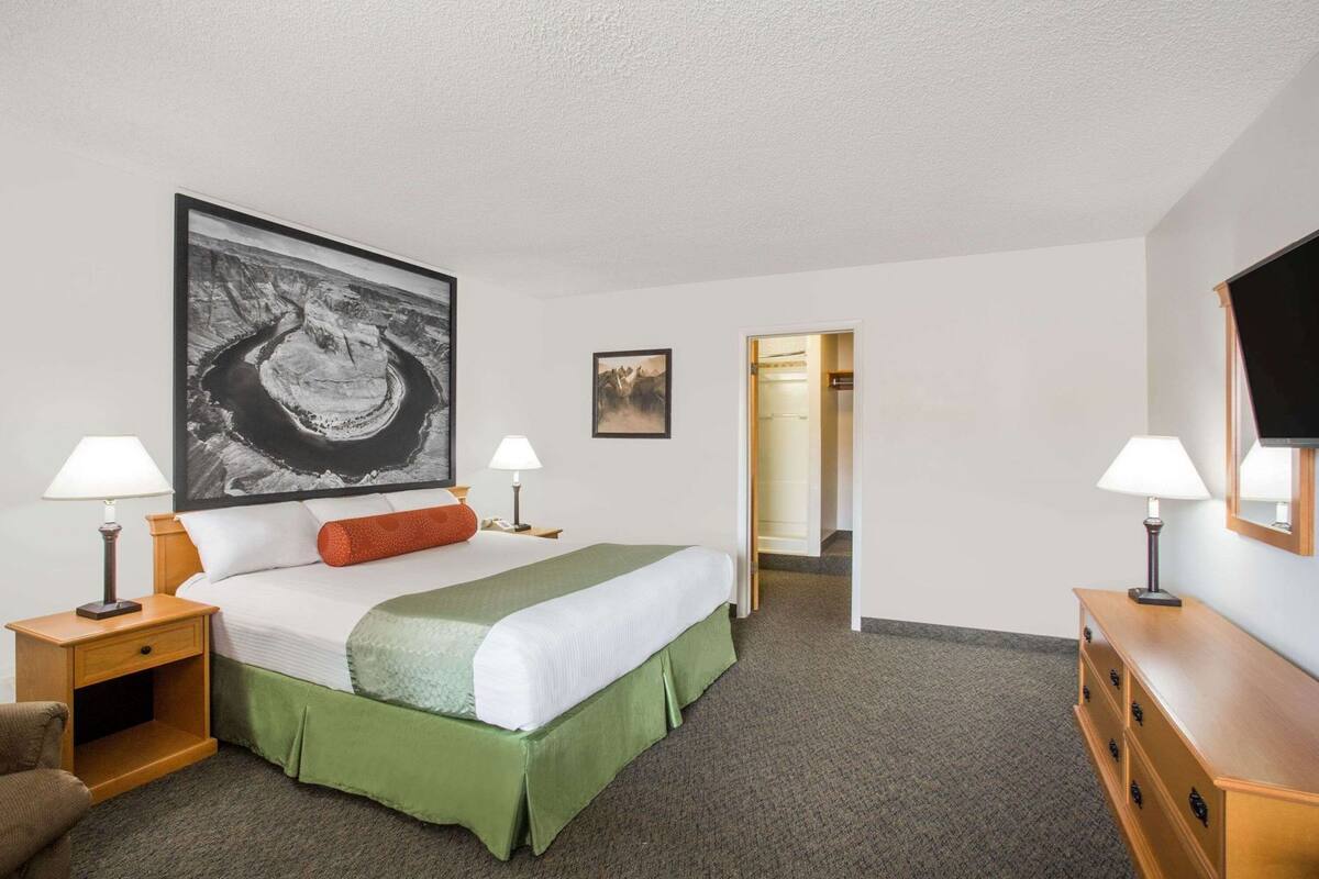 A hotel room with a bed, two bedside tables with lamps, a large framed picture above the bed, a television on the wall, and a door leading to a bathroom.