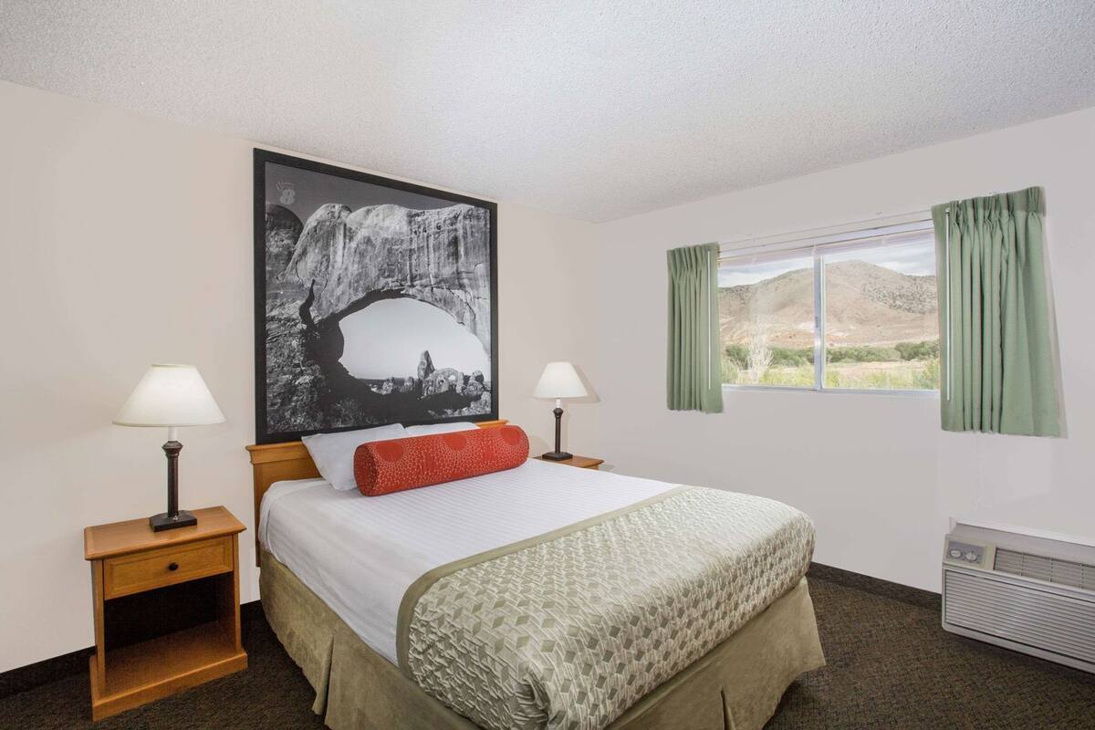 A hotel room with a bed, two bedside tables with lamps, a large painting on the wall, a window with curtains, and a radiator.