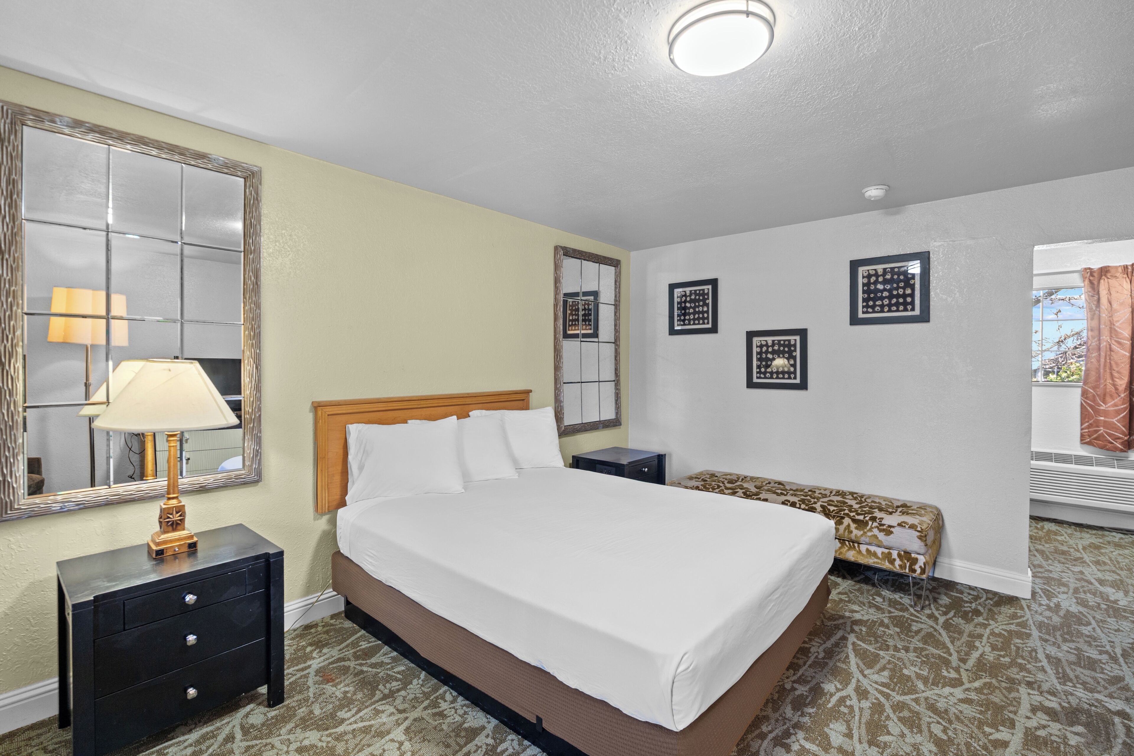 A hotel room with a bed, a nightstand with a lamp, a bench, and wall-mounted picture frames.