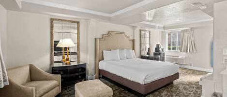 Habitación de hotel con una cama grande, una zona de estar con una silla y un reposapiés, un aparador, un espejo y una ventana con cortinas.