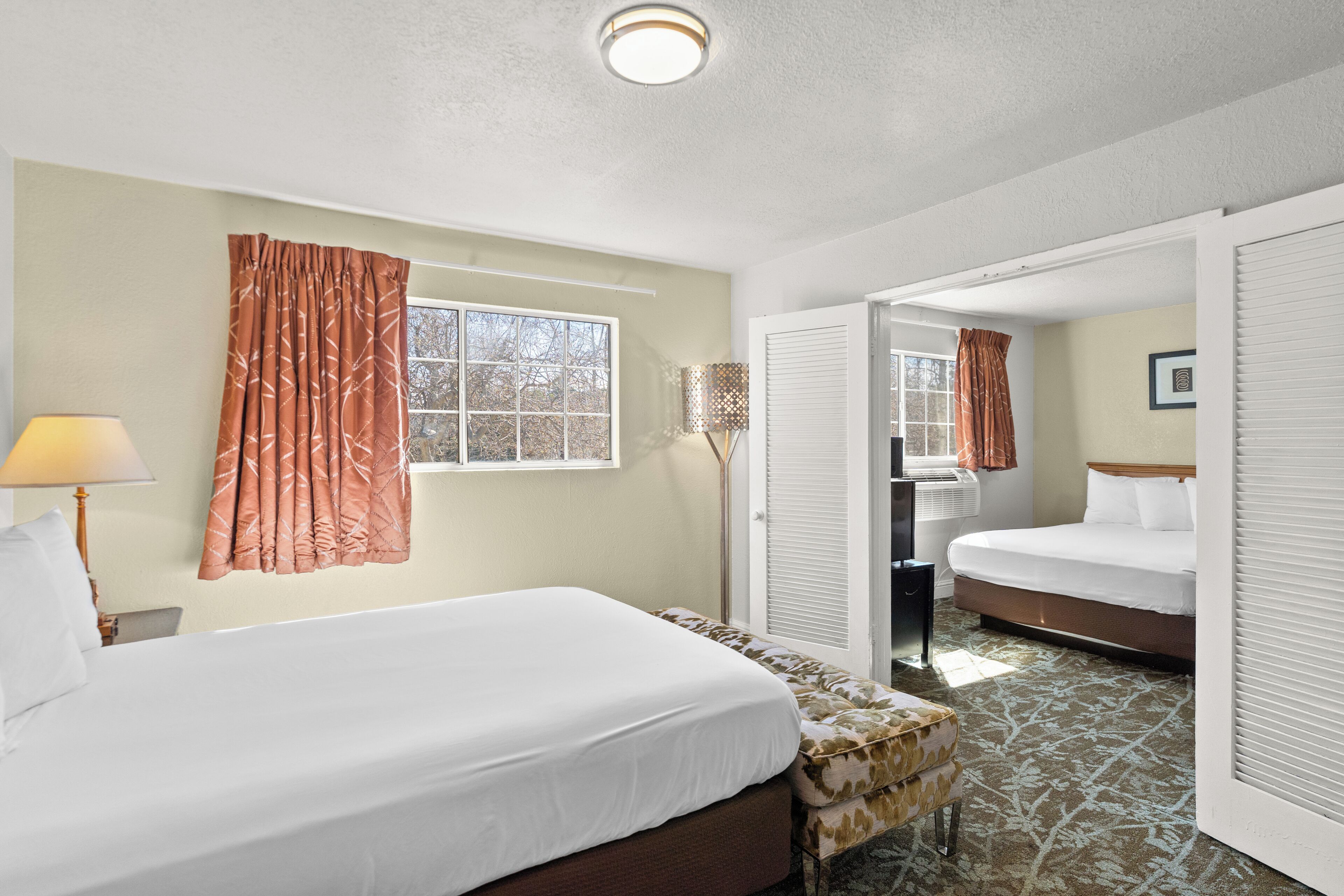 A hotel room with two beds, a window with curtains, a lamp, and a patterned rug.