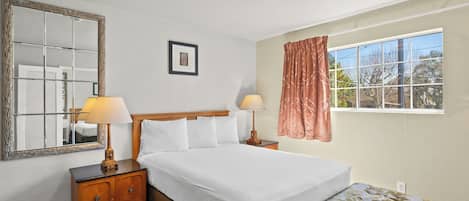 A hotel room with a large bed, two bedside tables with lamps, a mirror, a window with curtains, and a carpeted floor.