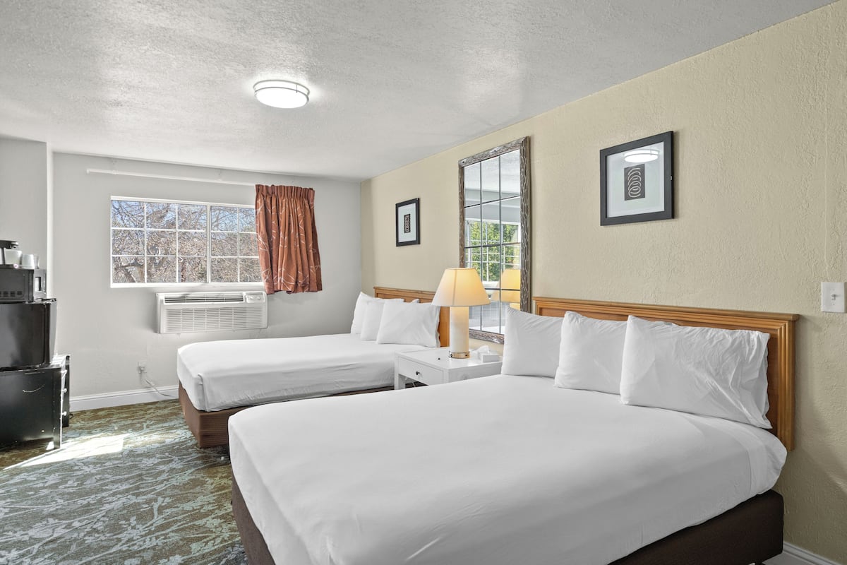 A hotel room with two beds, a window with curtains, an air conditioning unit, a bedside table with a lamp, and a framed picture on the wall.