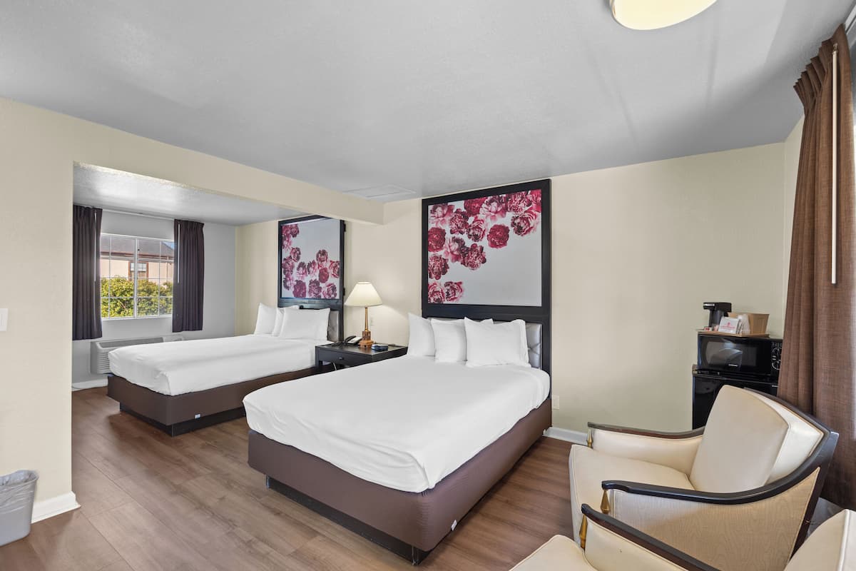 A hotel room with two beds, a chair, a desk with a lamp, and artwork on the walls.