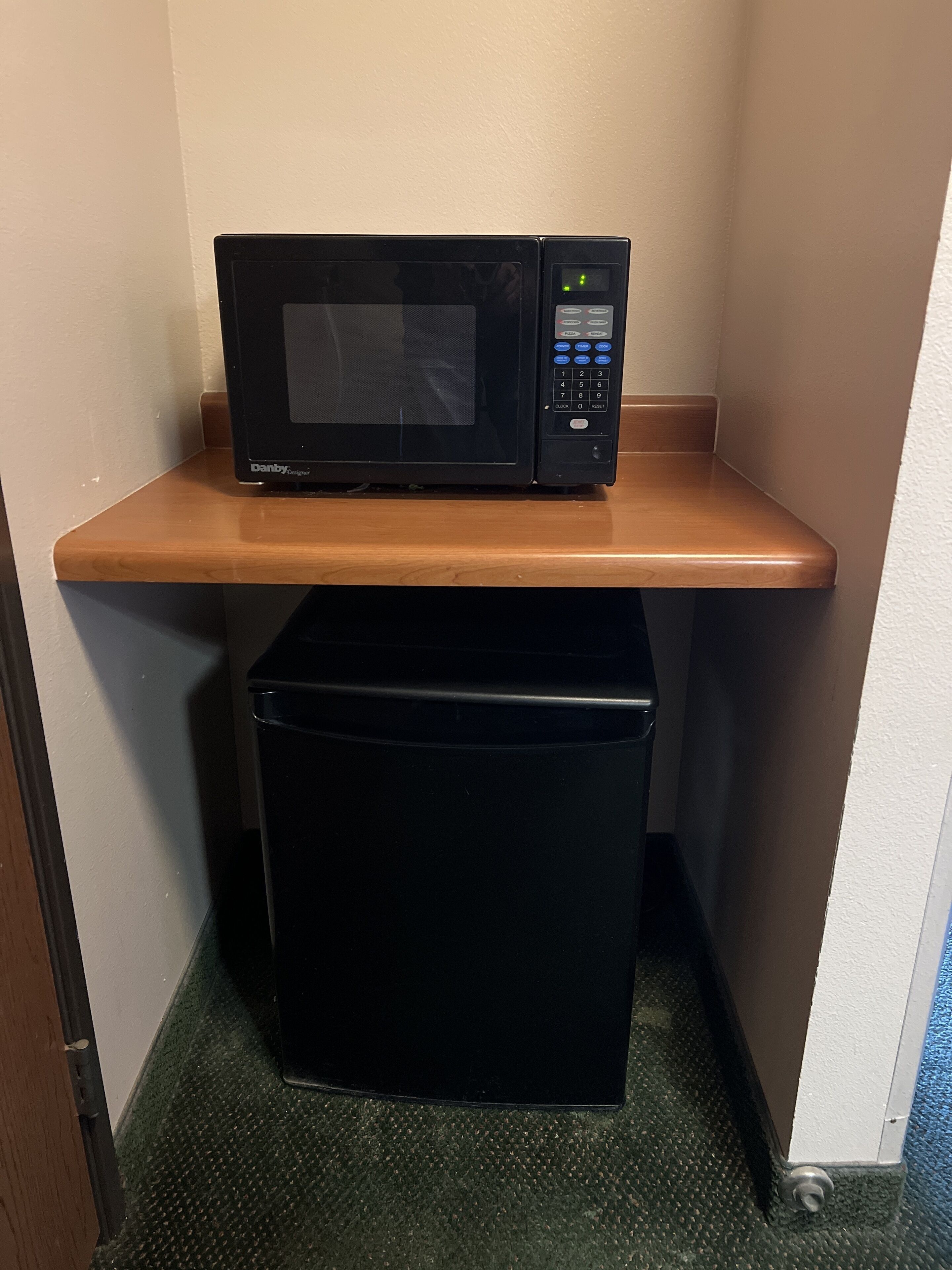 A black microwave on a wooden shelf in a corner of a room with a green carpet.