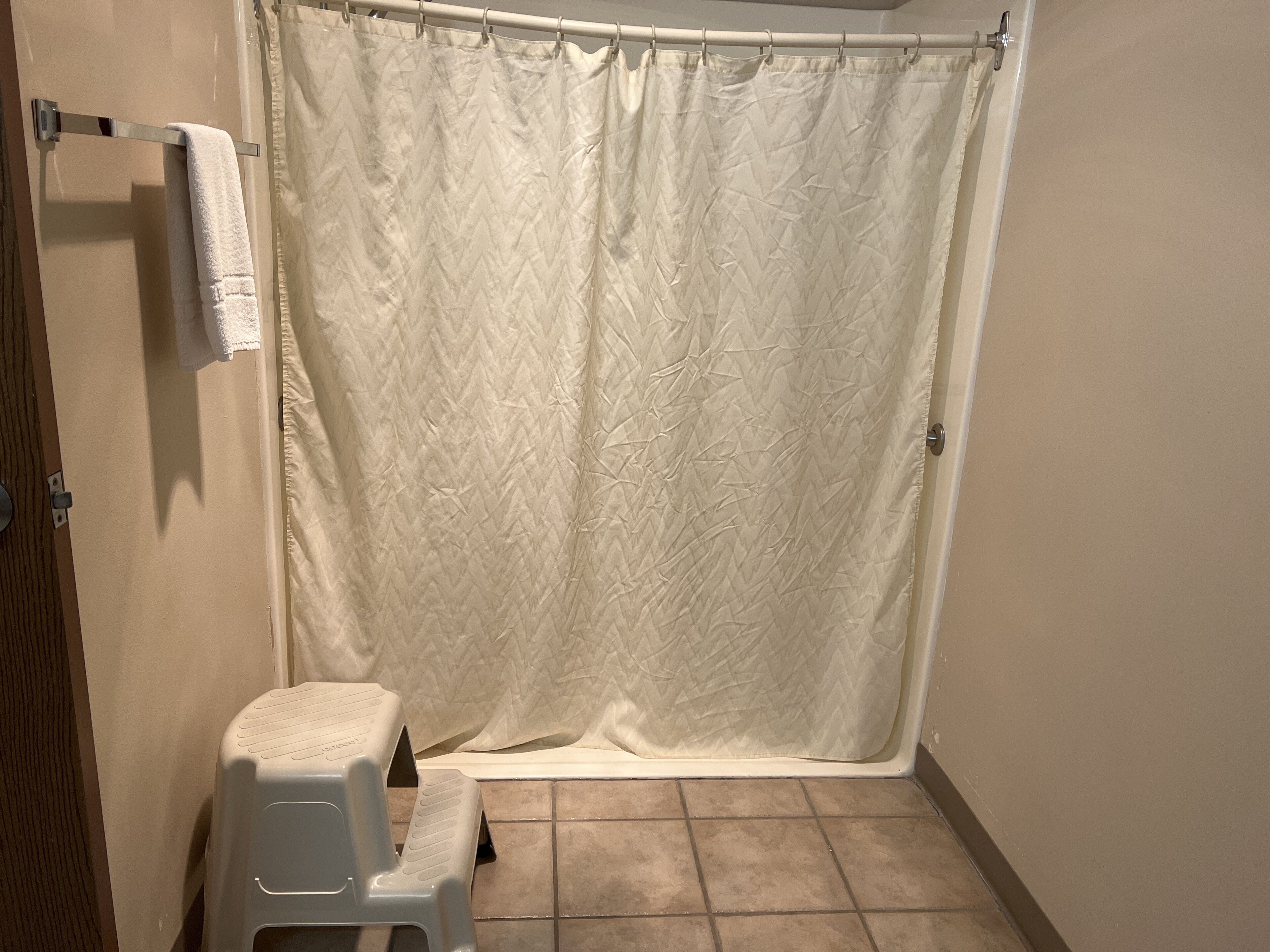 A bathroom with a shower curtain, a white plastic stool, and tiled flooring.