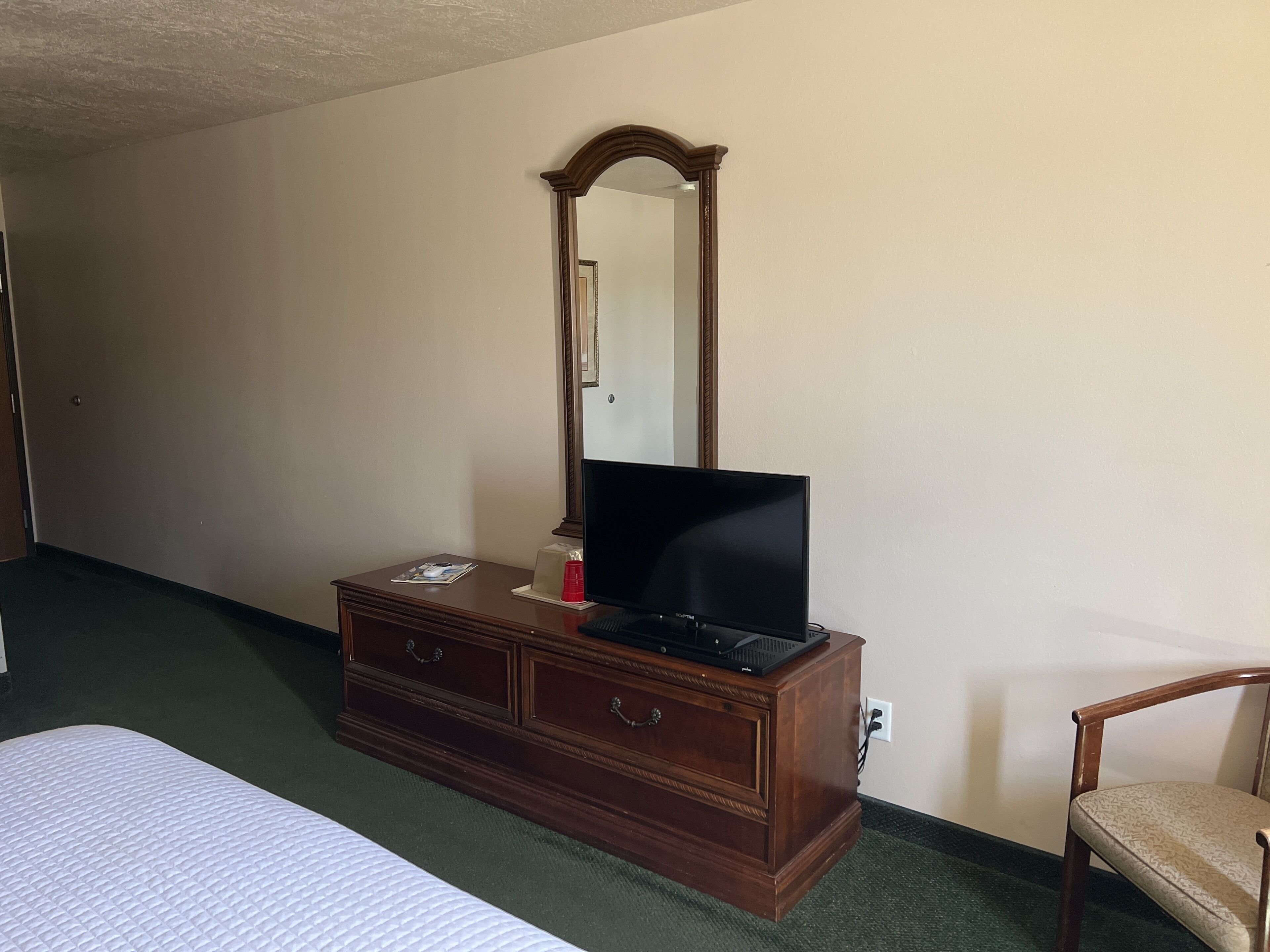 A hotel room with a wooden dresser, a flat-screen TV, a mirror, a bed, and a chair.