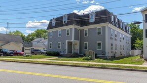A two-story residential building with a gray exterior, a front porch, and a driveway with parked cars.